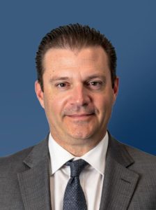 Chad Schumacher in a dark suit and tie against a blue background, bringing analytics leadership and data-driven insights across SAS and Kellogg's.