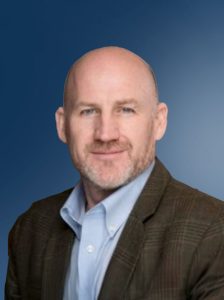 Dan Maxwell headshot with Mike Gamage in a brown blazer and light blue dress shirt against a smooth blue background with a bald look and trimmed goatee.