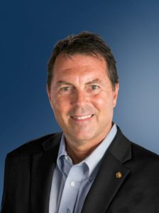 Smiling portrait of Rob-Haddock in a dark suit and light blue dress shirt against a clean blue studio backdrop.